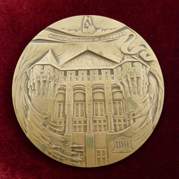 Medal owalny w kolorze złotym.Pośrodku rysunek bryły Pałacu Wielkich Mistrzów od strony wschodniej. Na dole logo UNESCO. Nad rysunkiem napis Muzeum Zamkowe w Malborku.