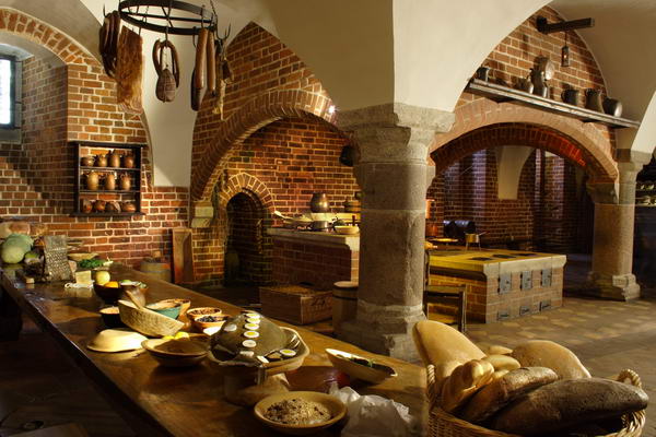 Pomieszczenie kuchni z ogromnym murowanym paleniskiem i okapem wspartym na dwóch kolumnach. Na ścianach półki z naczyniami Z lewej strony długi stół z dekoracją imitującą produkty spożywcze t. chleby, ryby, sery, mięsa i wędliny.