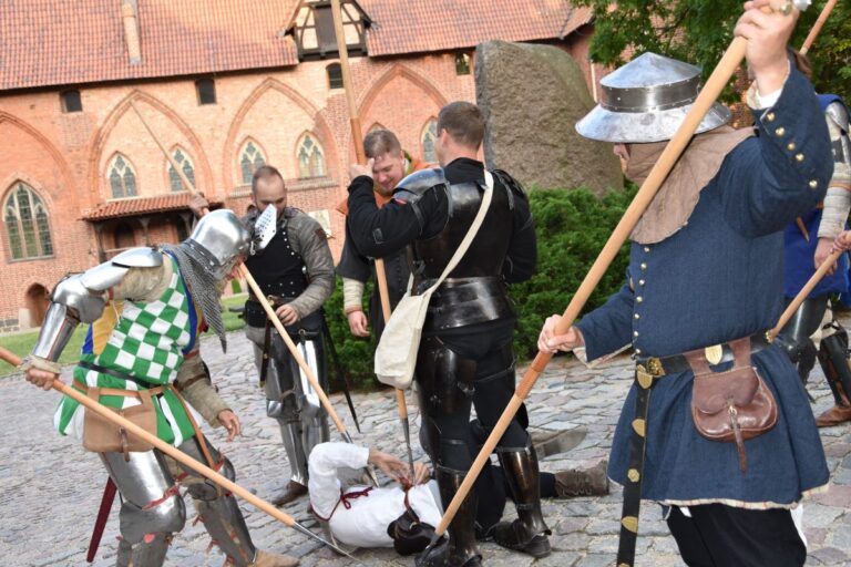 Dziedziniec Zamku Średniego. Sześciu mężczyzn w strojach z epoki stoi wokół leżącego mężczyzny . Pięciu z nich trzyma w rękach broń drzewcową markując uderzenia w leżącego.