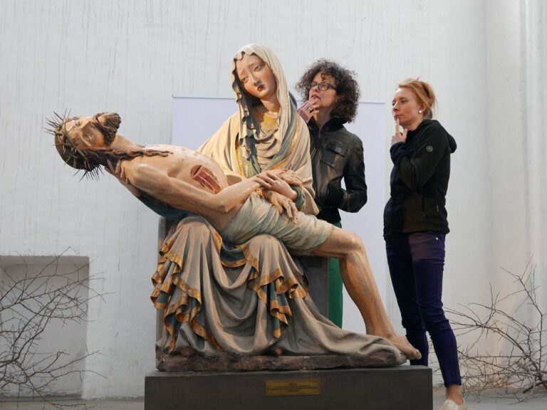 pośrodku na postumencie ustawiona rzeźba przedstawiająca Maryję trzymającą na kolanach martwego Chrystusa. Za obiektem stoją dwie kobiety oglądające dzieło.