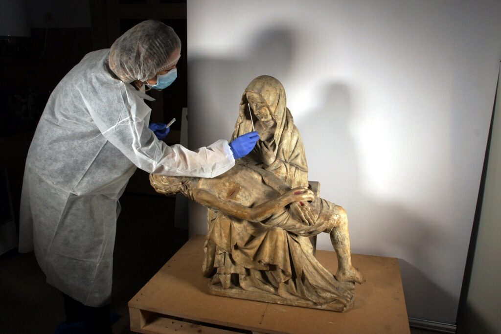 pośrodku na blacie stoi drewniana rzeźba przedstawiająca Maryję trzymającą na kolanach martwe ciało Chrystusa. obok stoi konserwatorka wykonująca prace przy obiekcie.