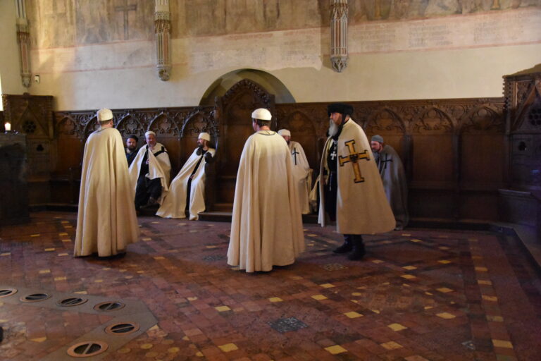 kilku mężczyzn w strojach średniowiecznych rycerzy zakonnych we wnętrzu kapitularza.