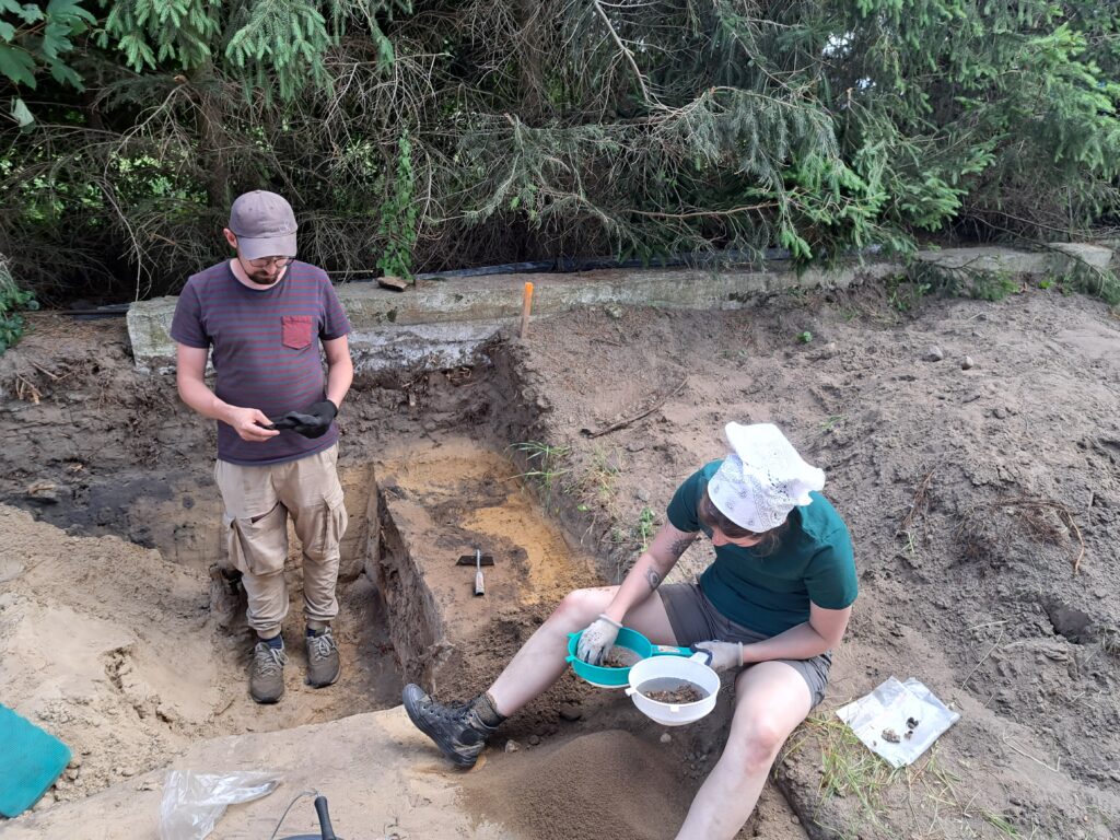 Archeolodzy podczas prac. W ziemnym wykopie dwie osoby. Mężczyzna stoi a w dłoniach trzyma ciemny niewielki przedmiot. Kobieta siedzi na krawędzi wykopu. W rękach ma dwa sitka, na których przesiewa ziemię.