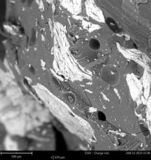 Zdjęcie próbki spod mikroskopu. kolorystyka czarno biała. Nieregularne kształty na całej powierzchni poniżej skala i dane liczbowe.