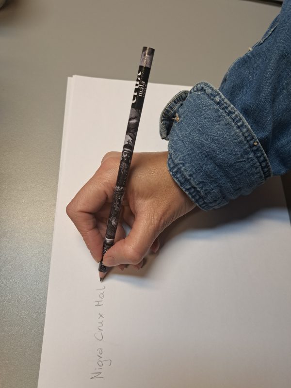 Kobieca dłoń trzyma czarny ołówek z logo wystawy i na kartce pisze Nigra crux