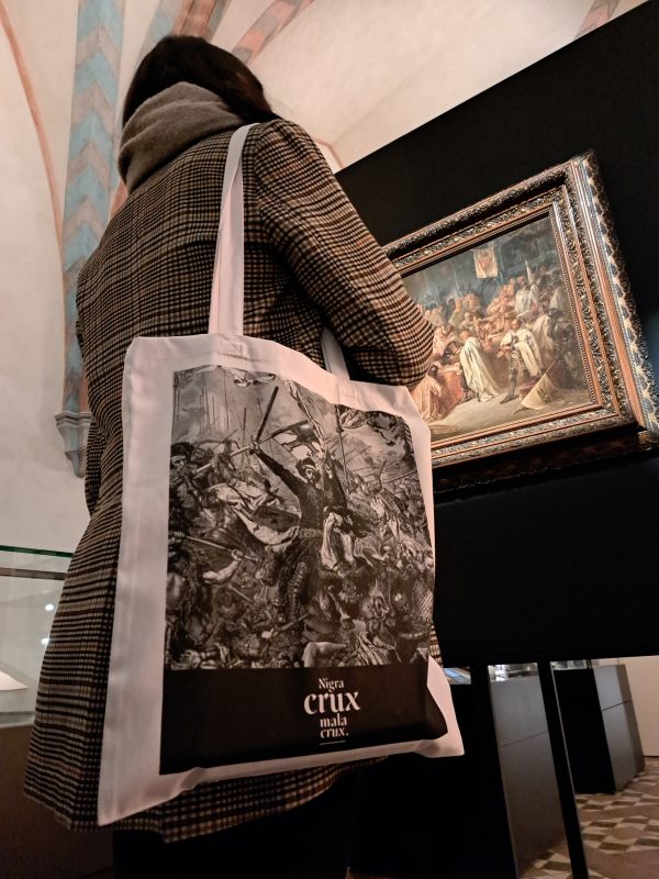 Przed obrazem na wystawie stoi kobieta. Na ramieniu ma płócienną białą torbę z odwzorowanym fragmentem z obrazu Jana Matejki Bitwa pod Grunwaldem. Na czarnym pasku na dole torby napis Nigra crux mala crux.