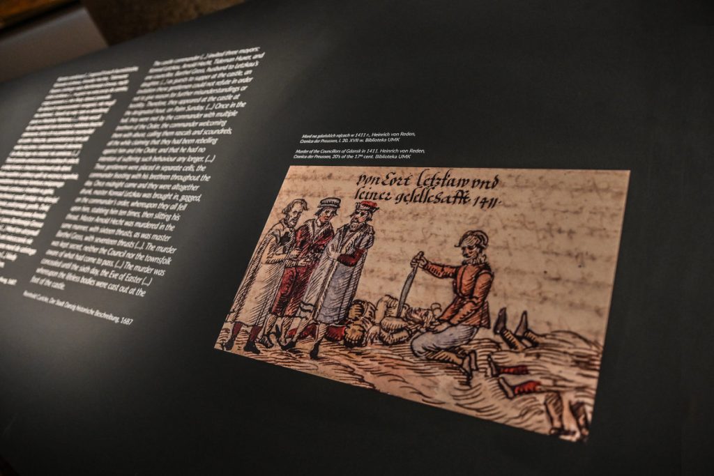 Czarny pulpit. Po lewej stronie tekst informacyjny, po prawej powiększona miniatura przedstawiająca czterech mężczyzn. Trzech stoi po lewej stronie, czwarty klęczy na ziemi pomiędzy ciałami martwych ludzi.