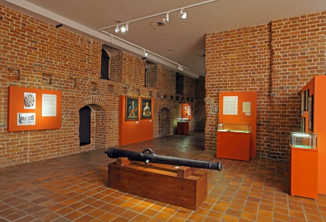 Exhibition hall. Brick walls with information boards and photos hung up. Exhibits in glass cases. In the middle, on a wooden pedestal, there is a bombard.