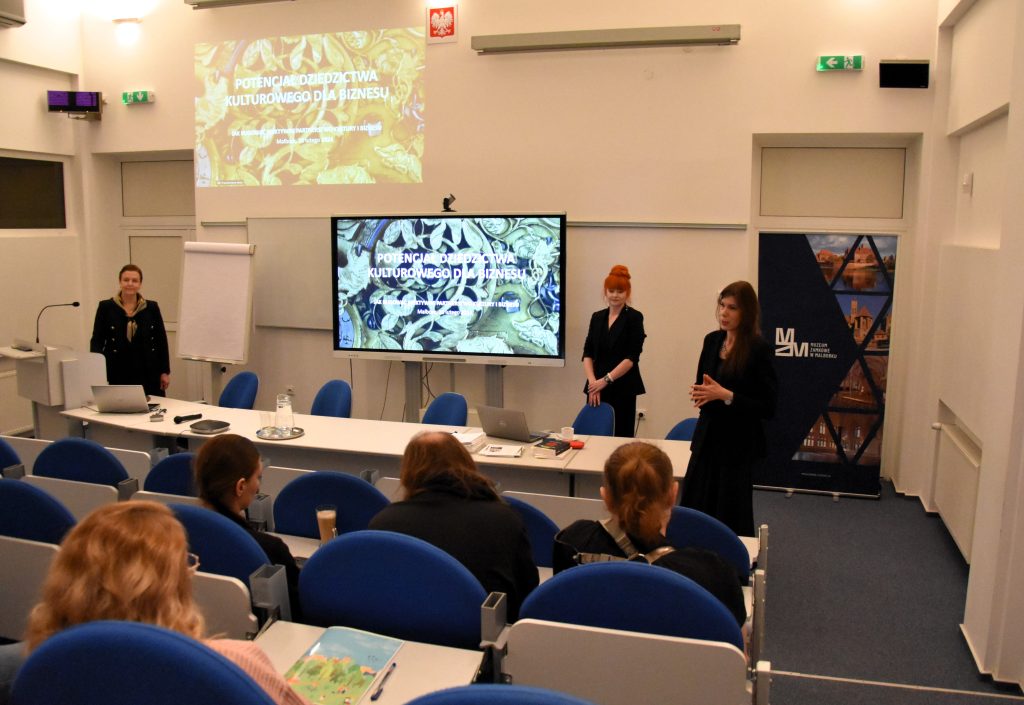 Sala konferencyjna. Ujęcie pokazuje od tylu siedzących uczestników. Na wprost obiektywu stół prezydialny, nad nim ekran z wyświetlaną prezentacją. Przy stole przodem do widowni trzy prelegentki.