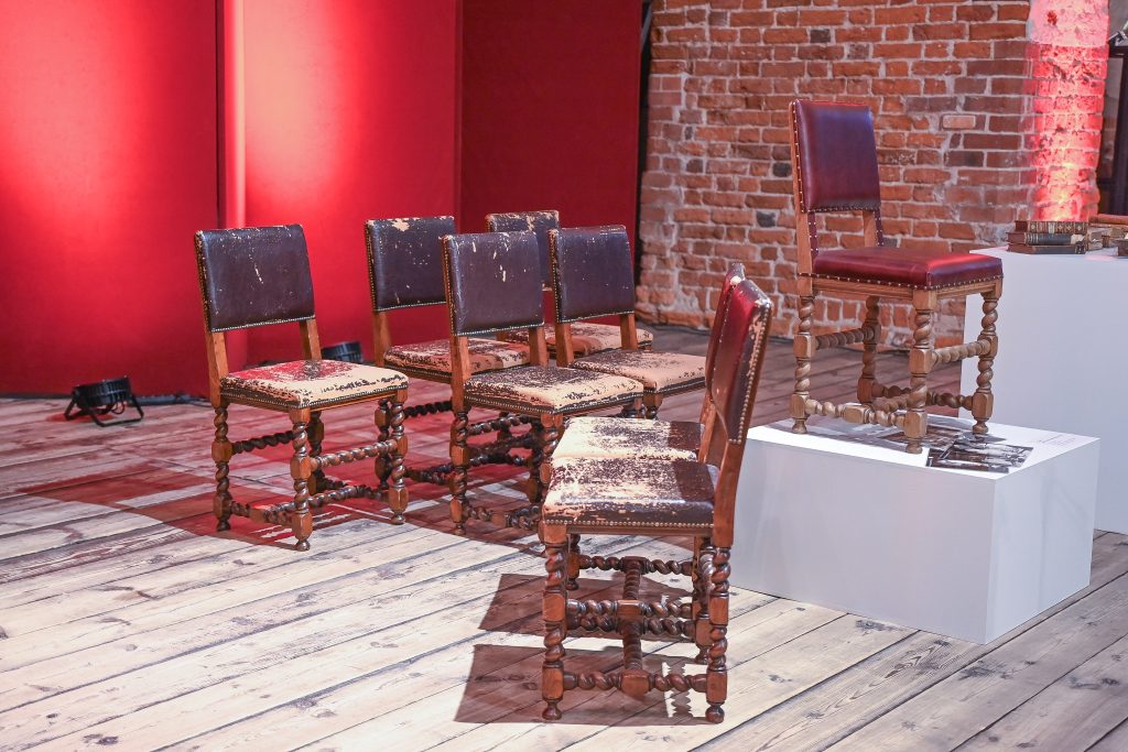 Na tle czerwonego parawanu i ceglanej ściany osiem krzeseł w tym jedno na podwyższeniu. Krzesła mają dekoracyjnie toczone nogi. Obite materiałem przypominającym skórę. Na całej ich powierzchni widoczne rozległe przetarcia.