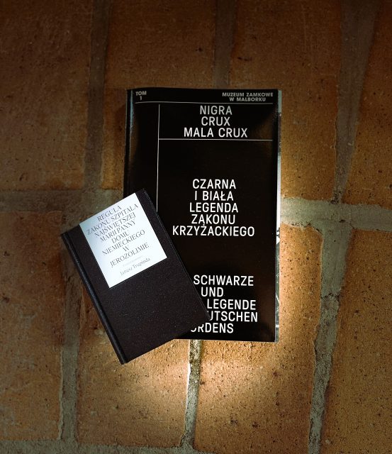 Na tle czerwonych cegieł leży duża książka w czarnej okładce z napisem Nigra crux mala crux. Czarna i biała legenda Zakonu Krzyżackiego. Na niej lekko po skosie nie zasłaniając tytułu położona druga dużo mniejsza książka w czarnej okładce z napisem na białym tle Reguła Zakonu Szpitala Najświętszej Marii Panny Domu Niemieckiego w Jerozolimie.