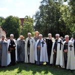 Na tle zielonych drzew przodem do widza stoi kilku mężczyzn ubranych w stylizowane na krzyżackie strojach.