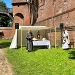 Fosa zamkowa. Po prawej stronie widoczny mur. Powyżej na wprost fragment drewnianego mostu prowadzącego na zamek. Przy murze ustawiony ołtarz polowy, nad nim baldachim. Z boku ołtarza stoi ksiądz. Przed ołtarzem stoi mężczyzna ubrany w szary habit.