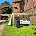 Fosa zamkowa. Po prawej stronie widoczny mur. Powyżej na wprost fragment drewnianego mostu prowadzącego na zamek. Przy murze ustawiony ołtarz polowy, nad nim baldachim. Przed ołtarzem stoi kobieta ubrana w stylizowany na średniowieczny strój w rękach trzyma otwartą książkę.