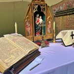 Ołtarz polowy. Na białym obrusie pośrodku drewniany ołtarzyk z Matką Bożą z dzieciątkiem. Po prawej stronie przybory liturgiczne. Po lewej na pulpicie otwarta księga z kartami zapisanymi odręcznym pismem.