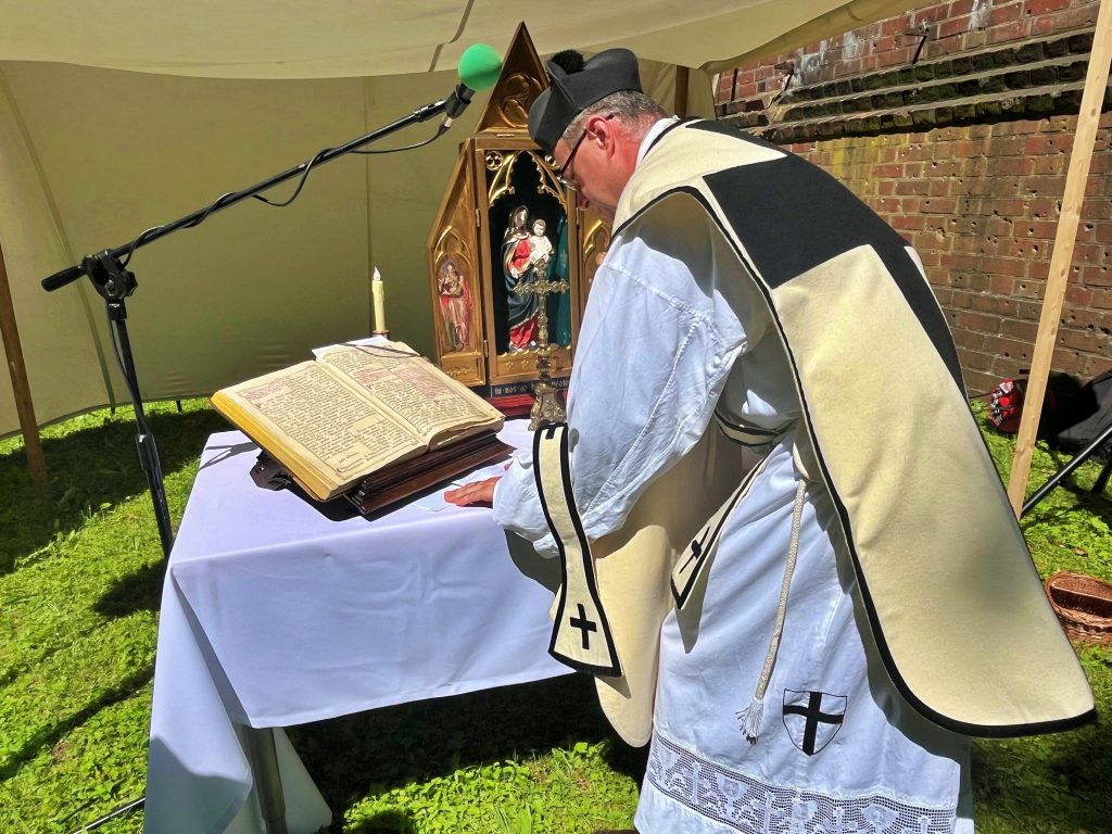 Ksiądz pochylający się nad ołtarzem polowym. Na białym obrusie pośrodku drewniany ołtarzyk z Matką Bożą z dzieciątkiem. Po lewej na pulpicie otwarta księga z kartami zapisanymi odręcznym pismem.