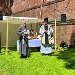 Ołtarz polowy w fosie zamkowej. Tyłem do ołtarza stoją ksiądz w białym ornacie z czarnym krzyżem i mężczyzna ubrany w strój stylizowany na średniowieczny habit.