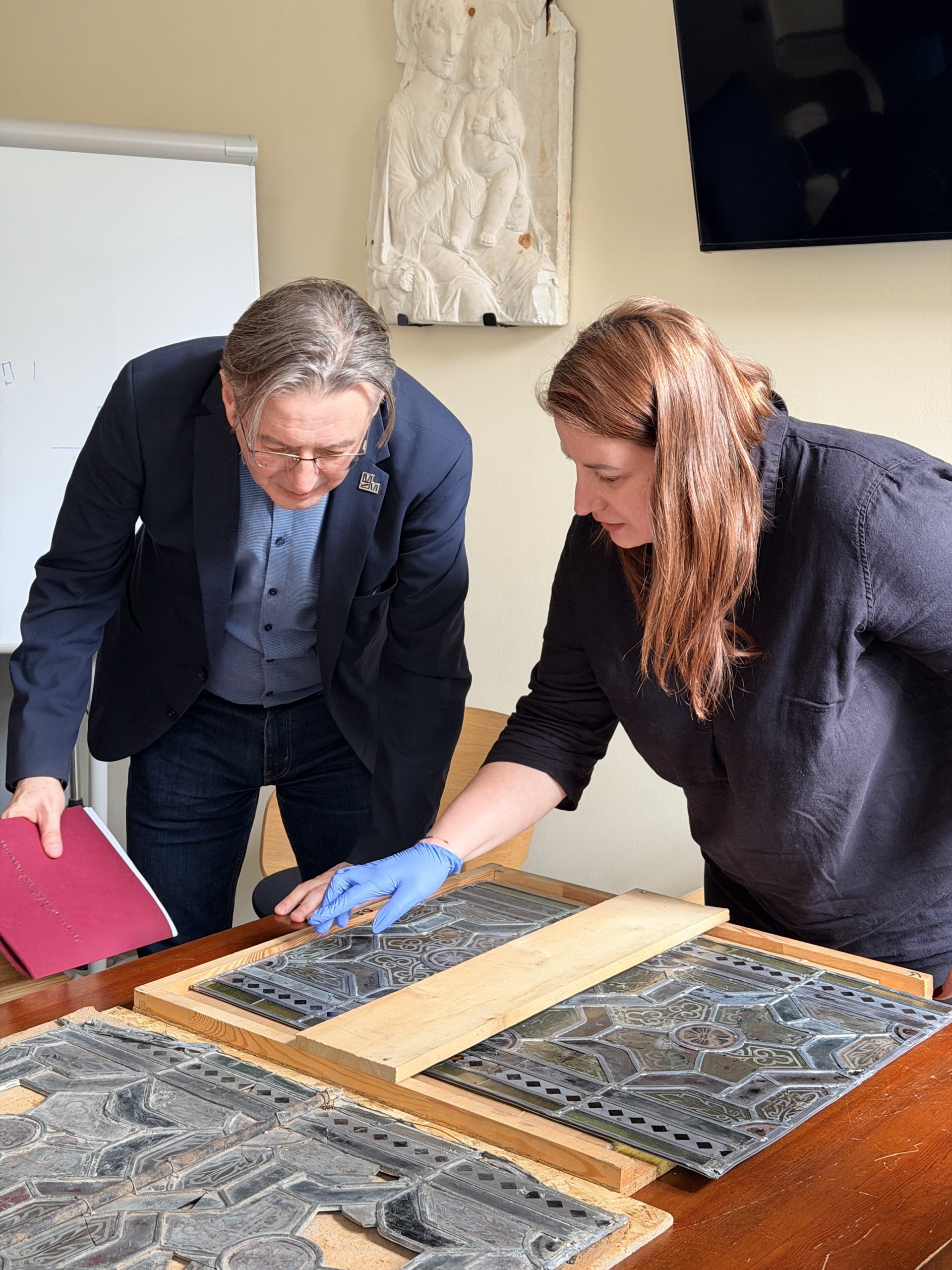 Two people leaning over a table examining decorative stained-glass panels; woman wears blue glove, man holds red folder.